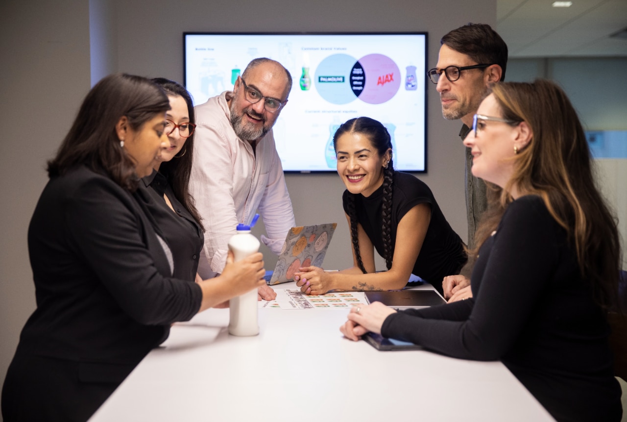 Six Colgate-Palmolive employees in a meeting room discussing a product concept, with a presentation slide on a screen behind them.