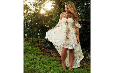 Artist Lani Balzan wearing a white lace high-low dress and a beaded feather necklace, standing in a lush green garden setting with the sun shining through the trees.